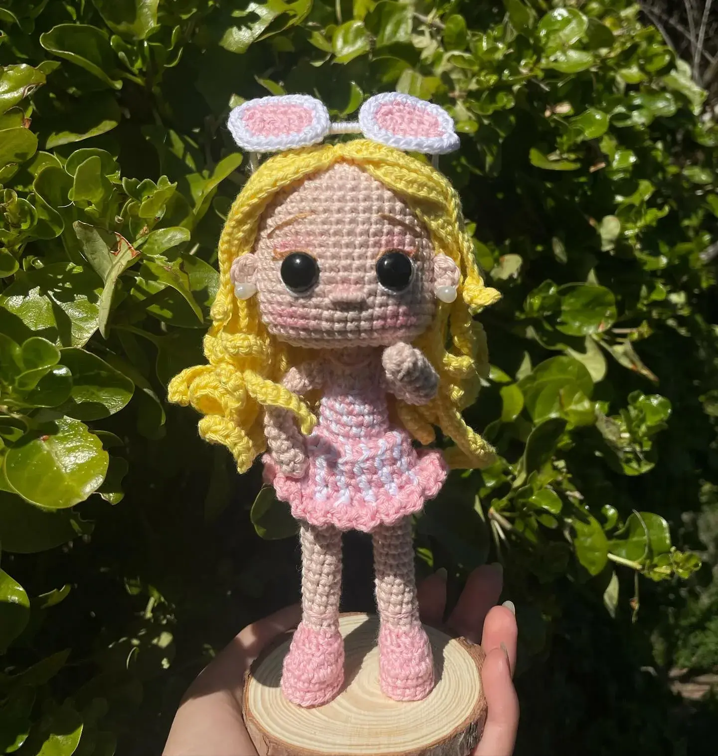 Muñeca amigurumi rubia con vestido rosado y lentes sobre la cabeza, en una base de madera